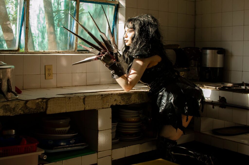 Cosplay character in dramatic lighting looking out of a kitchen window with scissor hands.