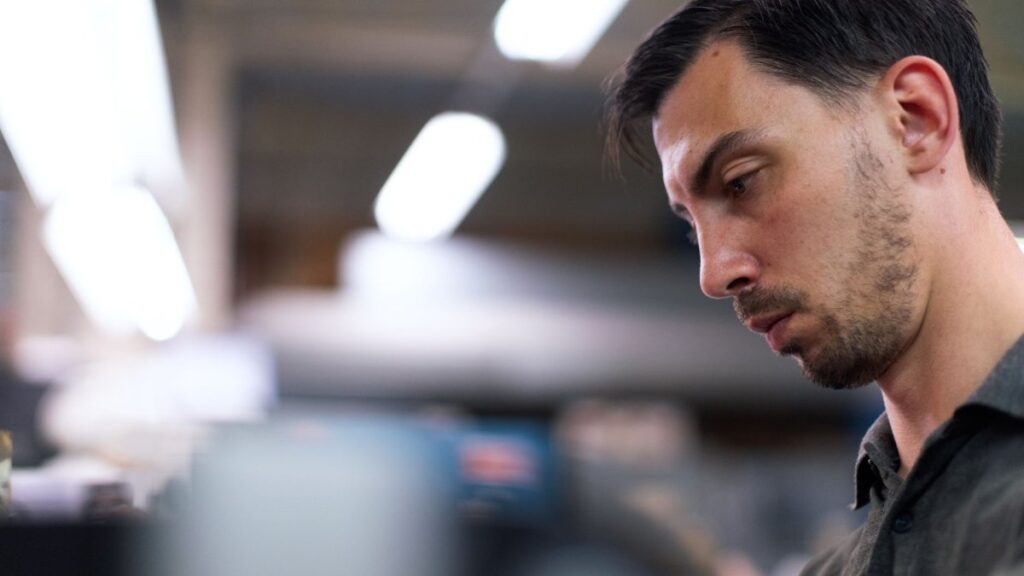 A man in a workshop focuses intently, surrounded by blurred tools and equipment.