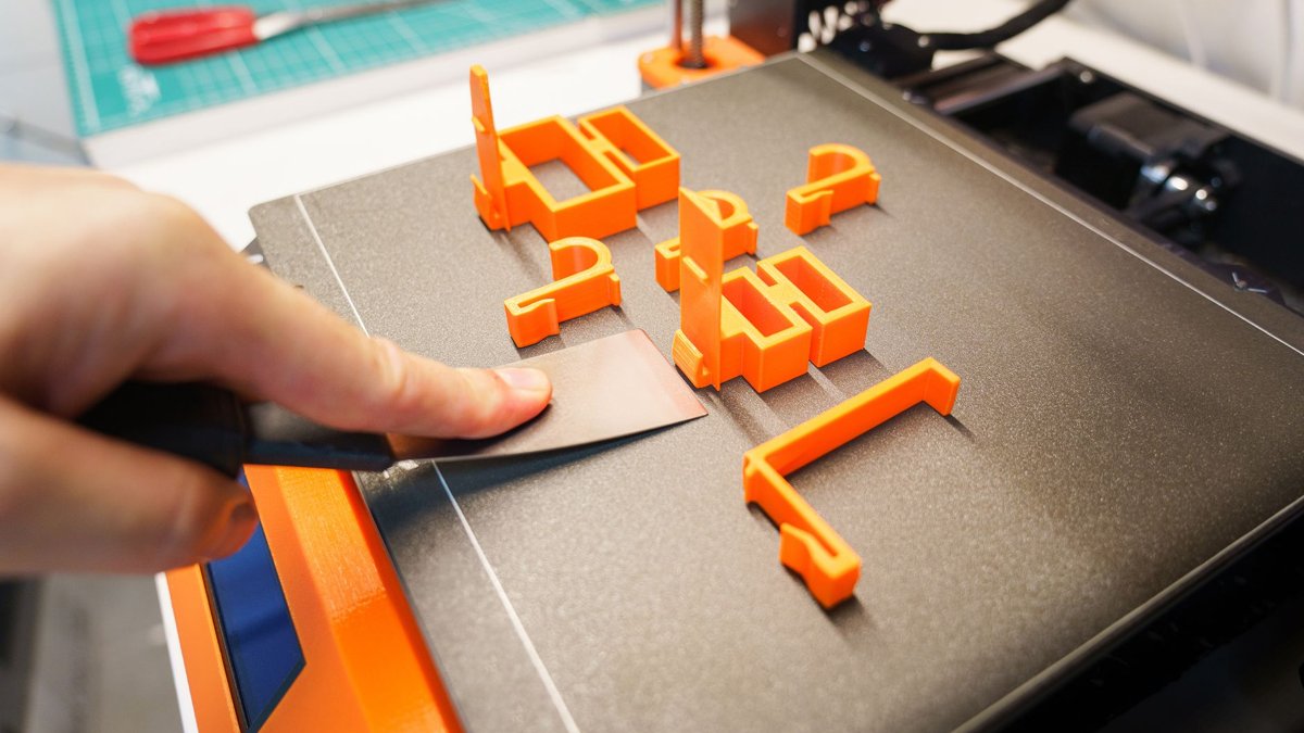 A hand carefully removes an orange 3D printed object from a printer bed using a spatula indoors.